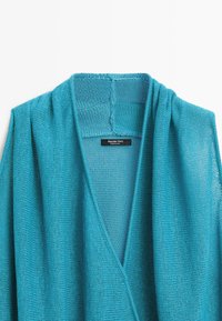 Cardigan en maille léger turquoise avec col châle et étiquette de marque visible à la couture du col.