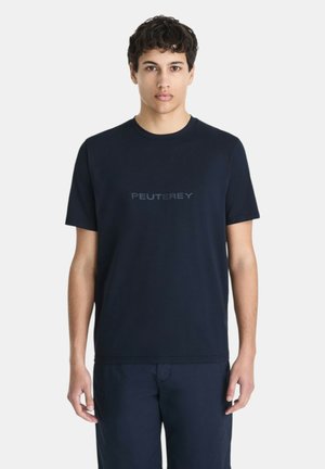 Jonge man met kort donker haar, gekleed in een donkerblauw Peuterey T-shirt en bijpassende broek,staand tegen een effen witte achtergrond.