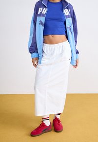 Person som bär en blå kort topp, en tvåfärgad blå Puma-jacka, en vit midi-kjol, vita strumpor med röda och svarta ränder, och röda sneakers.