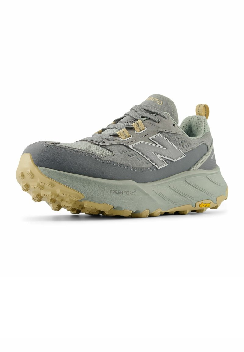 Chaussure de sport New Balance grise et beige avec une semelle épaisse, une tige en mesh, des lacets et une semelle texturée, inclinée pour montrer les détails avant et latéraux.