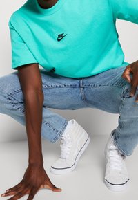 Tyrkysové bavlněné tričko s černým logem Nike, v kombinaci se světle modrými džíny a bílými teniskami vysokého střihu s černými akcenty.