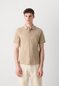 Polo beige en tissu doux, avec un col classique, des manches courtes et un petit logo sur la poitrine gauche. Porté avec un pantalon clair.