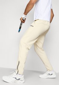 Pantalons de sport beige à coupe slim, avec un logo noir et des finitions noires contrastantes près de la cheville. Associés à des chaussures de sport blanches.