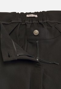 Black trousers featuring an elastic waistband, zip fly, snap closure, and pockets. Material appears smooth with minimal stitching details.