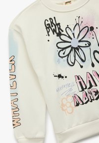 Sudadera crema con estampados gráficos en negro, rosa y azul. El diseño incluye una flor, elementos de texto y salpicaduras de pintura.