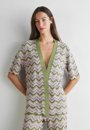 Femme portant un haut et un pantalon en maille assortis verts et lavande avec des manches trois-quarts, debout contre un mur blanc uni.
