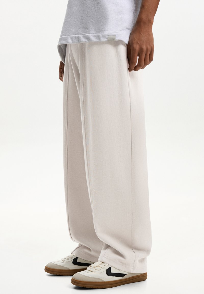 Light beige wide-leg pants made from soft fabric, featuring pleats, elastic waist, paired with cream and black sneakers.
