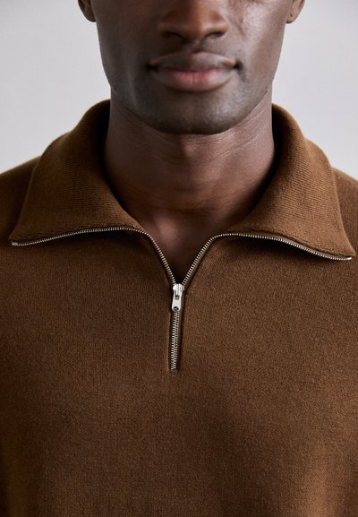 Gros plan sur une personne portant un pull en tricot marron avec une fermeture éclair argentée à demi-zip et un col replié.