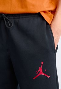 Nærbillede af en person iført sorte sweats og med et rødt Jumpman-logo og teksten "AIR" på låret samt en orange skjorte.