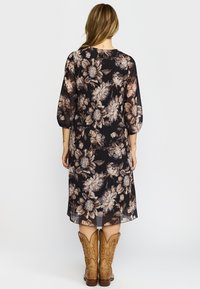 Kvinne med langt hår iført en svart midikjole med blomster og brune cowboy-støvler, stående med ryggen til mot en hvit bakgrunn.