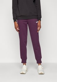 Lila träningsoverall med skräddarsydda fållar och en vit logotyp på låret, parat med vita sportskor. Svart sweatshirt bärs ovanpå.