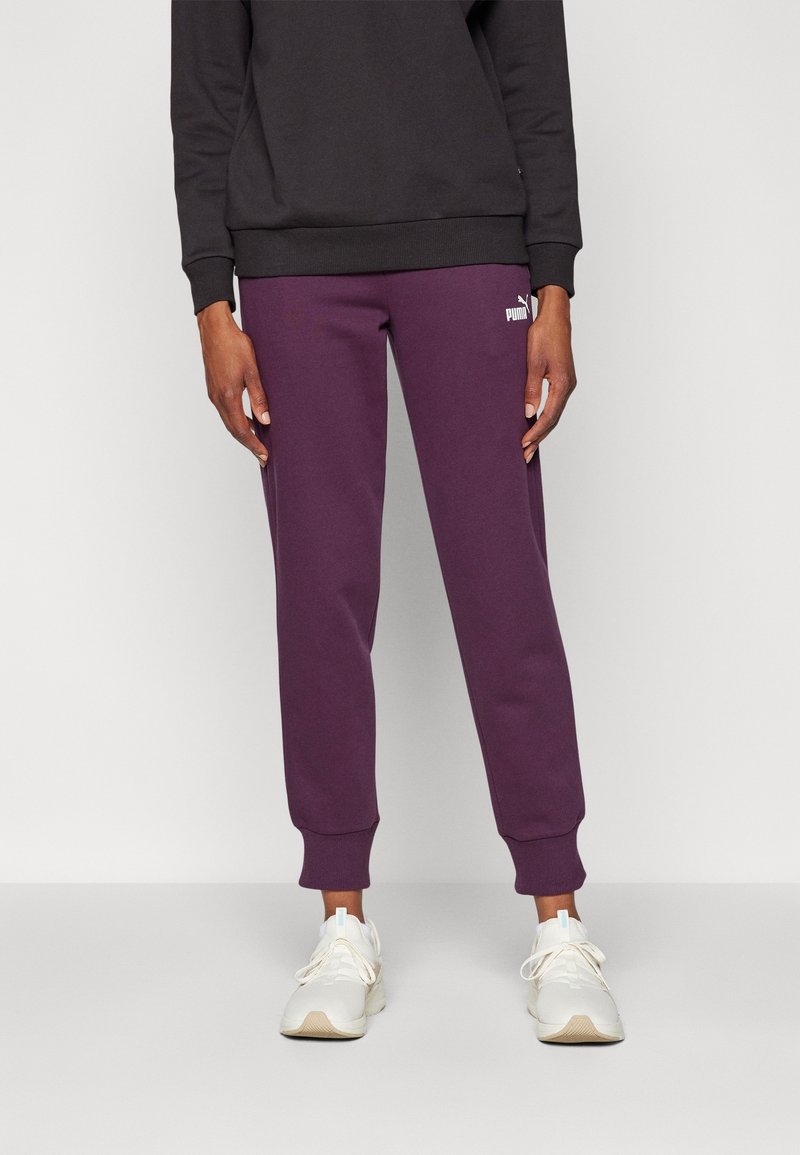 Lila träningsoverall med skräddarsydda fållar och en vit logotyp på låret, parat med vita sportskor. Svart sweatshirt bärs ovanpå.