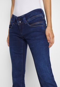 Tmavě modré džíny z denimu s přiléhavým střihem, vysokým pasem, zapínáním na dva knoflíky a kontrastními švy. Hladká textura.