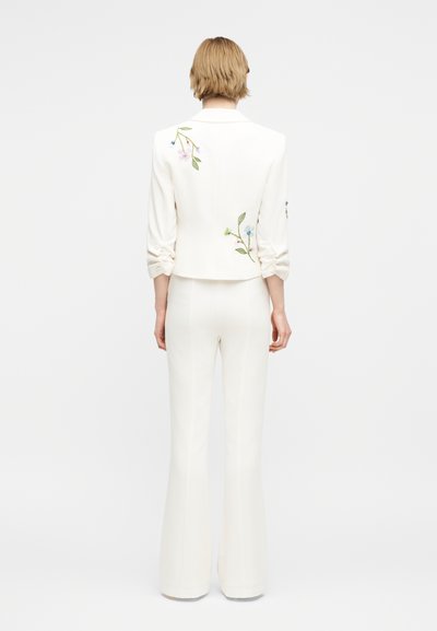 Άτομο με κοντά ξανθά μαλλιά που φοράει λευκή στολή με κεντήματα λουλουδιών στην πλάτη και τα μανίκια, στέκεται μπροστά σε απλό φόντο.