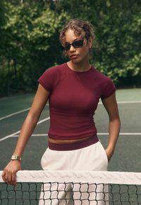 Mørkerød tætsiddende ribbet top med korte ærmer, parret med hvide sweatpants. Modellen bærer mørke solbriller og står ved et tennisnet.