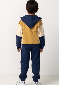 Kapuzensweatshirt in Senfgelb, Marineblau und Creme mit Farbblockdesign; kombiniert mit Marineblauen Jogginghosen. Das Material wirkt weich und strukturiert.