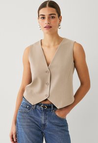 Beige ärmellose Weste mit tiefem V-Ausschnitt, drei Knöpfen vorne und kurzem Schnitt, kombiniert mit blauen Jeans.