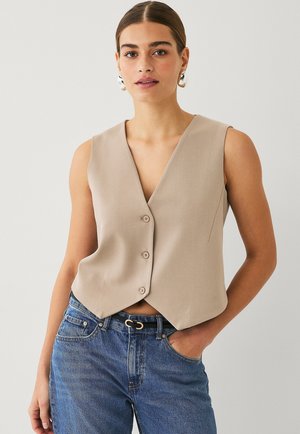 Beige mouwloos vest met een V-hals, voorzien van drie knopen aan de voorkant en een cropped ontwerp, gecombineerd met blauwe jeans. Gladde textuur.