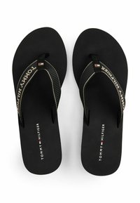 Chanclas negras con una tira texturizada que presenta el nombre de la marca en beige. La plantilla tiene un acabado mate y acentos con el logo.