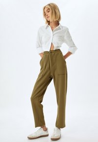 Olijfgroene losse broek met elastische tailleband en twee voorzakken, gecombineerd met een korte witte button-up blouse en witte sneakers.