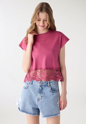 Giovane donna che indossa un top rosa con orlo in pizzo e shorts di jeans azzurri con toppe bianche in pizzo, in piedi davanti a uno sfondo semplice.