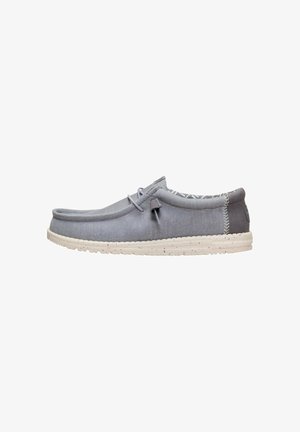 Zapato casual gris tipo slip-on con suela blanca moteada, parte superior suave, detalle de cordones en el lateral y collarín acolchado en el talón que muestra el interior con patrón.