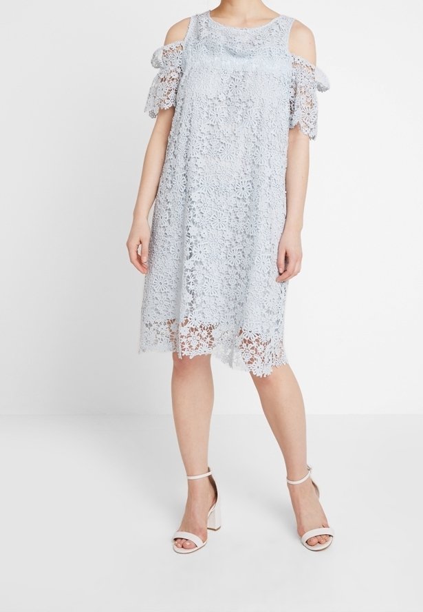 Femme portant une robe bleu clair à fleurs en dentelle épaules dénudées avec un ourlet festonné et des sandales blanches à talons blocs ouverts aux orteils, debout devant un fond blanc.