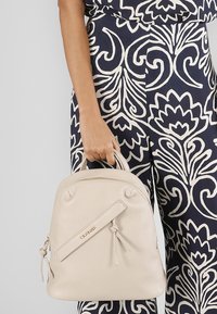 Beige kožená kabelka s zaobleným tvarem, horními uchy a dekorativním uzlovým detailem. Na přední straně se nachází jemné logo.