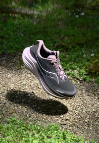 Zapatilla de running negra y rosa con parte superior de malla, cuello acolchado, suela texturizada y un logotipo distintivo en el lado, suspendida sobre una superficie natural.