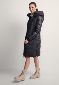 Kobieta w czarnej, sięgającej do kolan kurtce puchowej z kapturem i beżowych botkach na sznurowadła, stojąca na neutralnym tle, lekko się uśmiechająca.