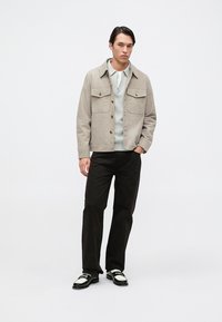 Giovane uomo in piedi, indossa una giacca beige con bottoni sopra un maglione grigio chiaro con zip, pantaloni neri e mocassini bianco e nero su sfondo bianco semplice.