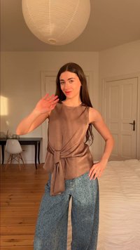 Jeune femme aux longs cheveux foncés faisant signe de la main, portant un haut marron sans manches et un jean large bleu, debout dans une pièce lumineuse avec un sol en bois.