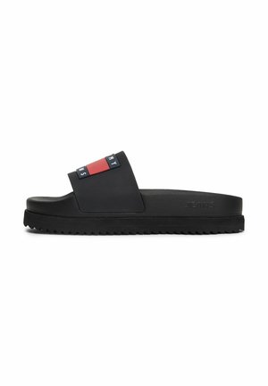 Schwarze Slide-Sandale mit dicker Sohle und rotem, weißem und blauem Logo-Riemen mit der Aufschrift "Tommy Jeans" auf weißem Hintergrund.