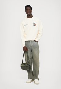 Homem vestindo uma sweatshirts creme com gráfico de cassete, jeans cinzentos largos, tênis brancos, segurando uma bolsa transversal verde ao lado, em pé sobre um fundo branco.