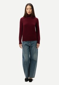 Femme debout portant un pull bordeaux à manches longues avec un col montant, un jean bleu à jambes larges et des chaussures noires pointues sur un fond uni.