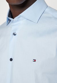 Mężczyzna ubrany w jasnoniebieską koszulę z guzikami i szerokim kołnierzykiem, z małym prostokątnym logo w kolorach czerwonym, białym i granatowym na piersi.