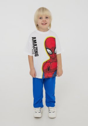 T-shirt bianco con una grafica colorata di Spider-Man, abbinata a pantaloni blu. Sneakers bianche con chiusure a velcro completano l'outfit.