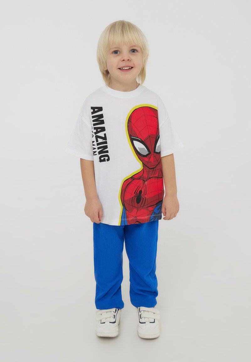 T-shirt bianco con una grafica colorata di Spider-Man, abbinata a pantaloni blu. Sneakers bianche con chiusure a velcro completano l'outfit.