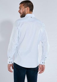 Weißes Langarmshirt mit klassischem Kragen, geknöpften Manschetten und glatter Textur. Besitzt einen kleinen schwarzen Akzent auf der Rückseite.