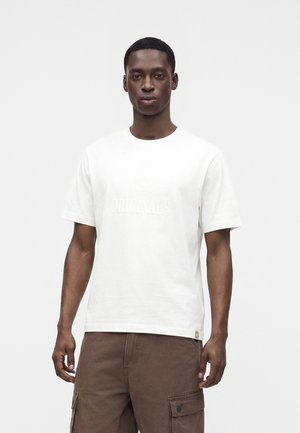 Homme portant un t-shirt blanc uni à manches courtes avec un texte subtil "ORIGINALS" et un pantalon cargo marron, debout devant un fond blanc.