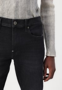 Jeans de denim oscuro con un diseño ajustado, que presentan un cierre de botón plateado y costuras sutiles en los bolsillos. Acompañados de un suéter de canalé gris claro.