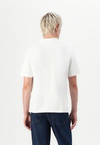 White polo shirt with short sleeves, open collar, and a smooth texture. Worn with blue jeans, viewed from the back against a plain background.