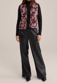 Väst med blommönster i rosa och svart över en svart topp, kombinerad med blanka svarta vidbyxor. Mjuka texturer och skräddarsydd passform.