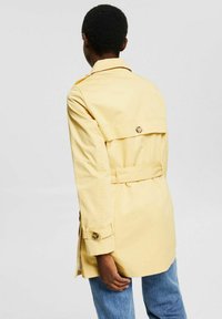 Trench-coat jaune avec une taille ceinturée, des épaulettes et des accents de boutons. Coupe ample avec manches longues et fente à l'arrière.