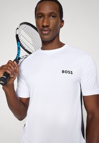 Λευκό αθλητικό μπλουζάκι με μαύρο λογότυπο, με raglan μανίκια και πλαϊνές λεπτομέρειες, κρατούμενο από ένα άτομο με ρακέτα τένις σε ουδέτερο φόντο.