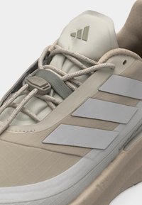 Adidaș bej cu trei dungi albe, șireturi elastice fără necesitatea de a fi legate, cu un dispozitiv de reglare gri, și limbă amortizată care prezintă un logo triunghiular.