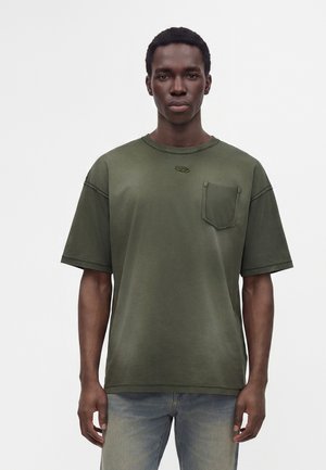 Jeune homme portant un t-shirt oversize vert olive avec une poche poitrine et un jean clair, debout devant un fond blanc uni.