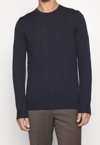 Man som bär en marinblå långärmad tröja med rund hals och bruna skräddarsydda byxor mot en enkelt ljus bakgrund.