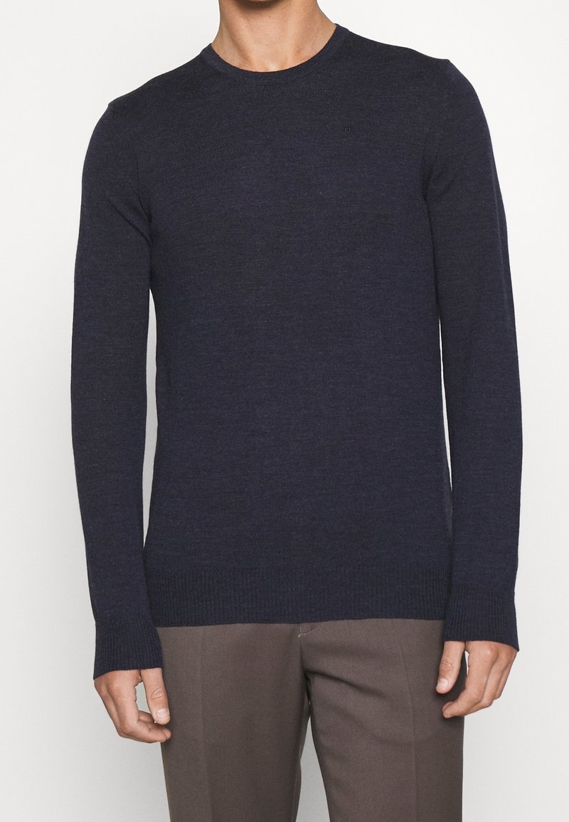 Man som bär en marinblå långärmad tröja med rund hals och bruna skräddarsydda byxor mot en enkelt ljus bakgrund.