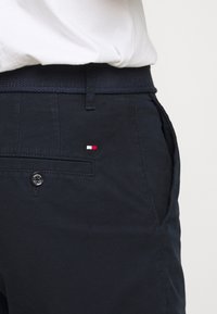 Námořnické modré kalhoty s texturovaným pásem, které mají malé logo v červené, bílé a modré barvě blízko kapsy a detail s knoflíkem.